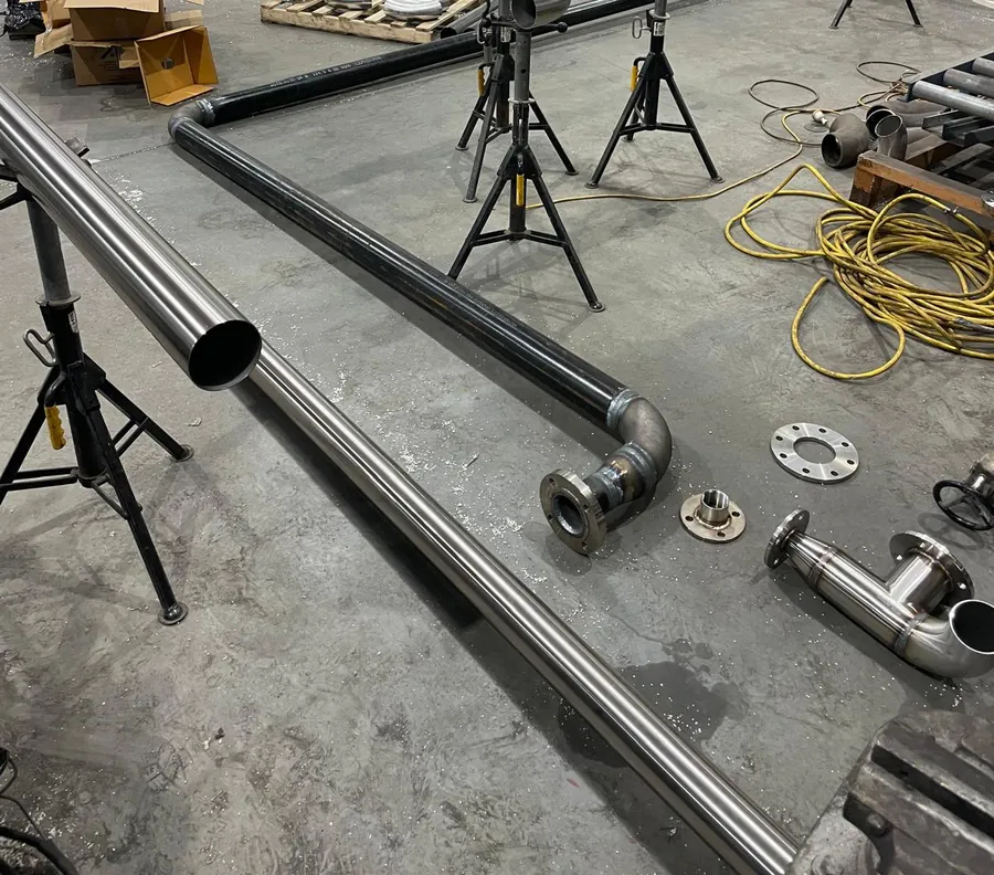 Stainless and black steel pipe with bends and fittings set up on pipe stands, with flanges laid out on a factory floor