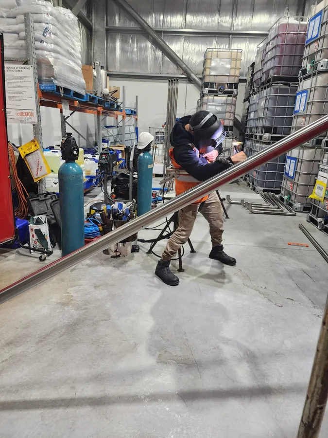 Welder in a welding helmet, hi-vis vest and gloves welding a long stainless steel tube in a warehouse
